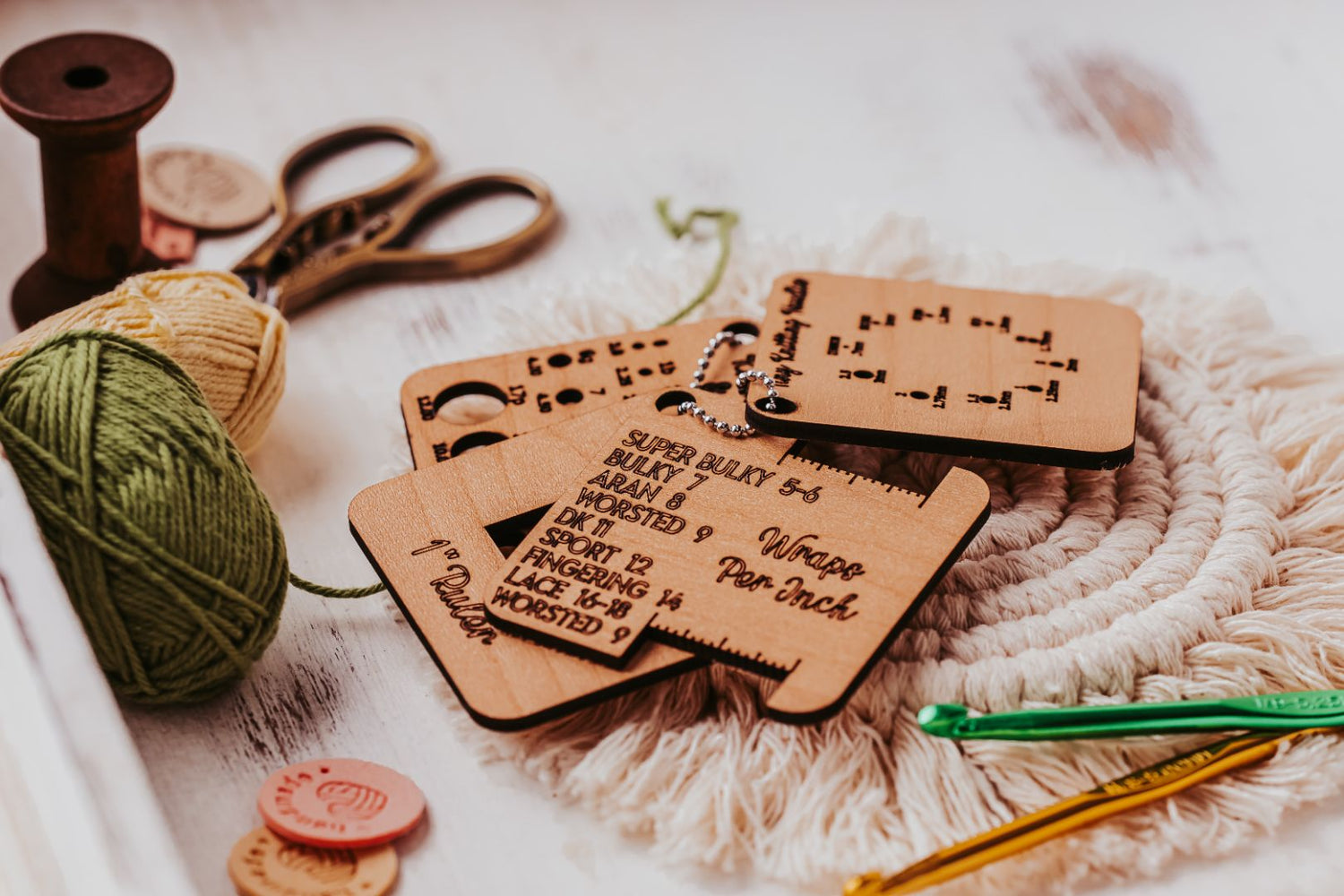 Wooden crochet gauge tools with etched measurements are laid on a woven mat. Nearby are crochet hooks, a spool of thread, green and yellow yarn, and scissors on a white surface.