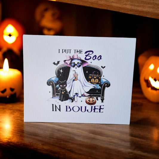 The Funny Halloween Sign from Mirabilia Boutique, titled "I put the Boo in Boujee," is displayed on a wooden surface, surrounded by lit jack-o'-lantern candles. The sign features a ghost wearing a pink witch hat and sunglasses, sitting on a couch with black cats and pumpkins. Perfect for any ghost enthusiast's Halloween decor!