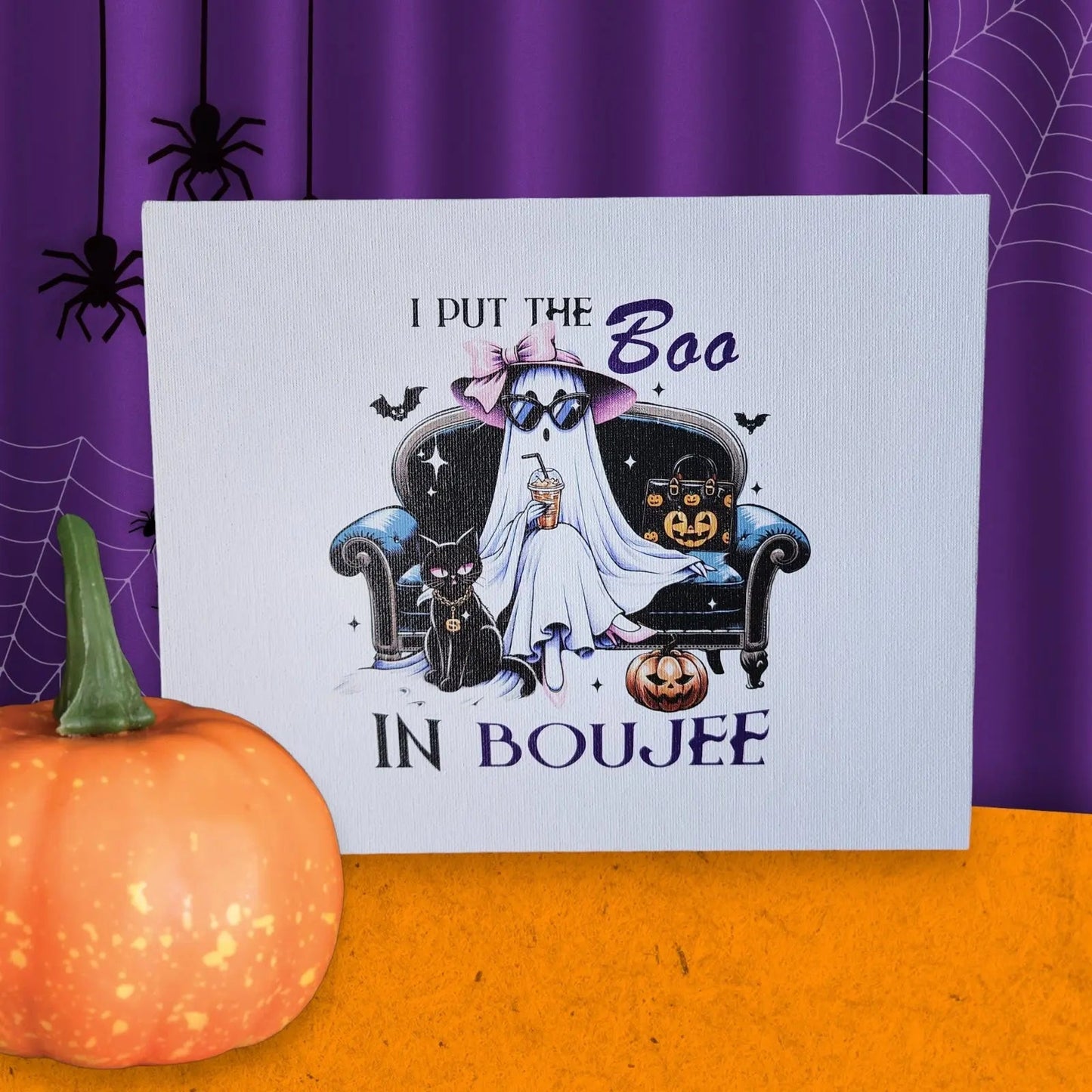 A Halloween-themed canvas panel sign from Mirabilia Boutique, titled "Funny Halloween Sign: I put the Boo in Boujee," is displayed on a table beside a small pumpkin. The sign portrays a ghost enthusiast in a fashionable outfit, relaxing on an elegant couch amidst pumpkins, bats, and a black cat. In the background hang purple curtains adorned with spiderweb designs.