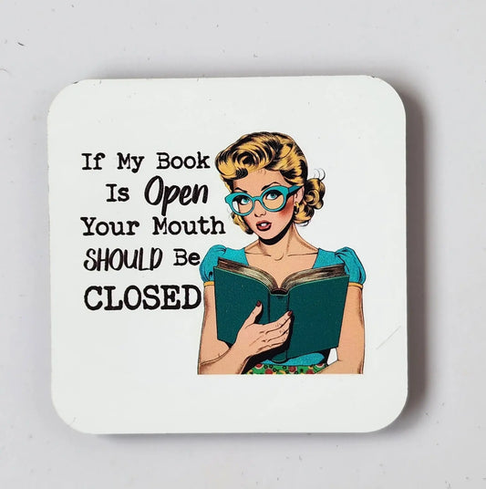 A cartoon illustration features a woman with blonde hair, blue glasses, and a teal top holding an open book. The accompanying text reads, "If my book is open, your mouth should be closed." This design is showcased on a square white coaster by Mirabilia Boutique—an ideal gift for any book lover that celebrates literary passion.