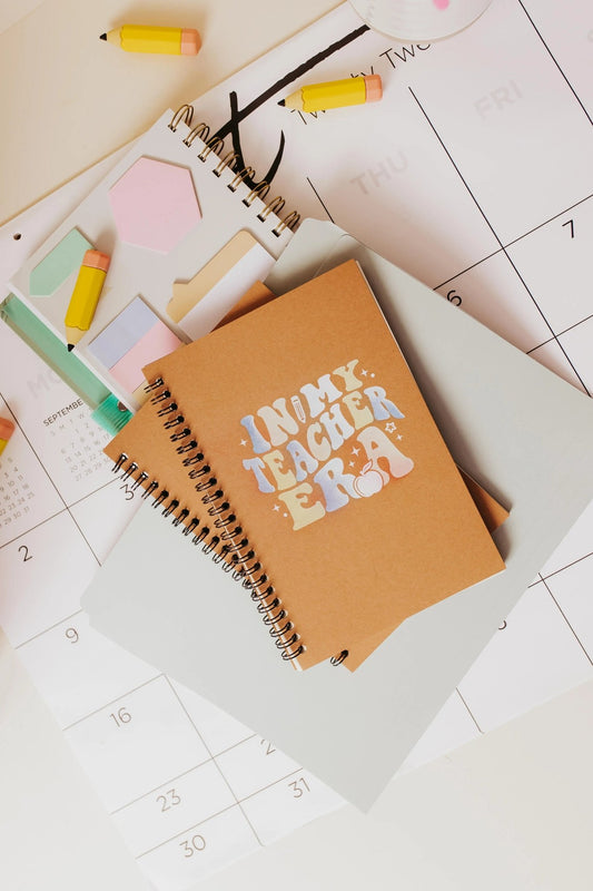 An "In My Teacher Era" notebook by Mirabilia Boutique, featuring a spiral-bound design, sits atop a calendar. Accompanying it are sticky notes, vibrant mini erasers, and a pencil, which together create a lively and organized workspace atmosphere—an ideal gift for educators.