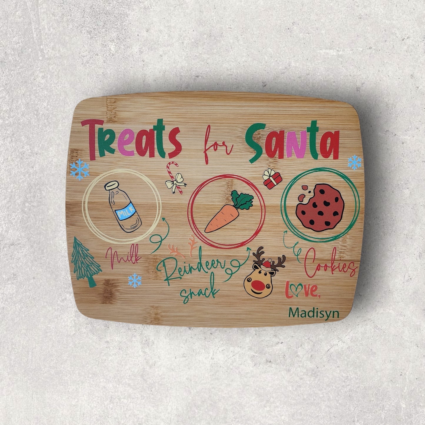 The Santa Cookie Board Personalized by Mirabilia Boutique features sections for milk, a carrot for reindeer, and cookies. Adorned with festive drawings of a Christmas tree, reindeer, heart, and gift, it embodies the Christmas Eve tradition with the label "Treats for Santa." This unique board is personalized with the name "Madisyn.