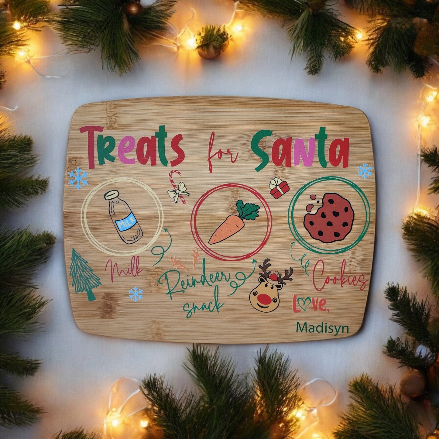 The "Santa Cookie Board Personalized" by Mirabilia Boutique is ideal for your Christmas Eve tradition. It features charming illustrations of a milk bottle, a carrot, and a cookie labeled "Milk," "Reindeer snack," and "Cookies," all surrounded by festive pine branches and string lights. Additionally, it can be personalized with the name “Madisyn” at the bottom.