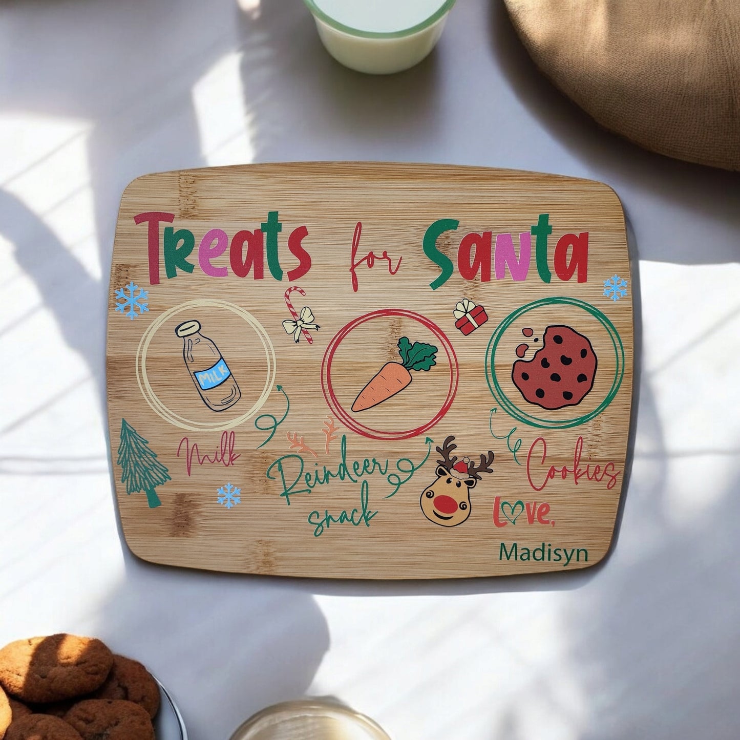 The Santa Cookie Board Personalized by Mirabilia Boutique is a charming Christmas Eve tradition featuring sections for milk, a carrot, and cookies. Adorned with colorful text and festive drawings of a tree, reindeer, and snowflakes, it includes the name "Madisyn" displayed at the bottom.