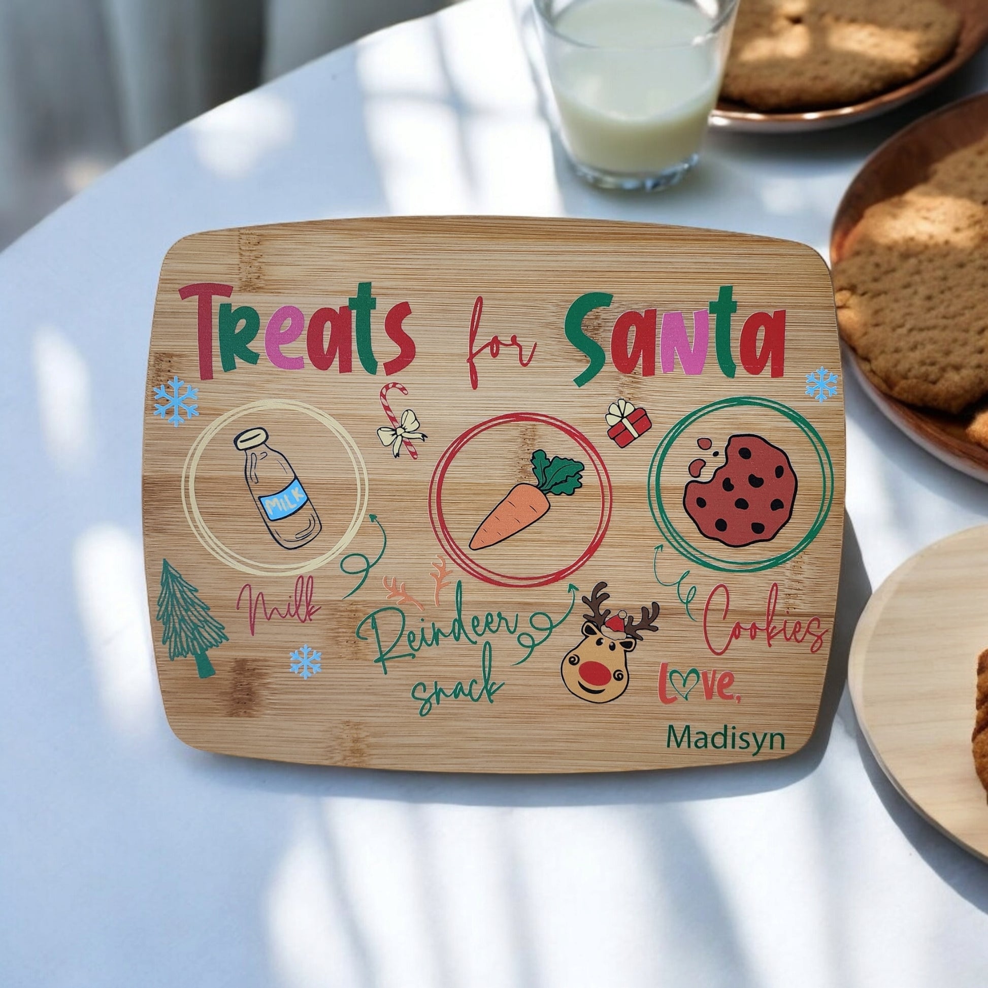 A Santa Cookie Board Personalized by Mirabilia Boutique is placed on the table, elegantly labeled "Treats for Santa." It includes designated areas for milk, a reindeer snack (carrot), and cookies. This festive board is decorated with charming holiday illustrations and complemented by a glass of milk and a plate of cookies, creating an ideal Christmas Eve tradition.