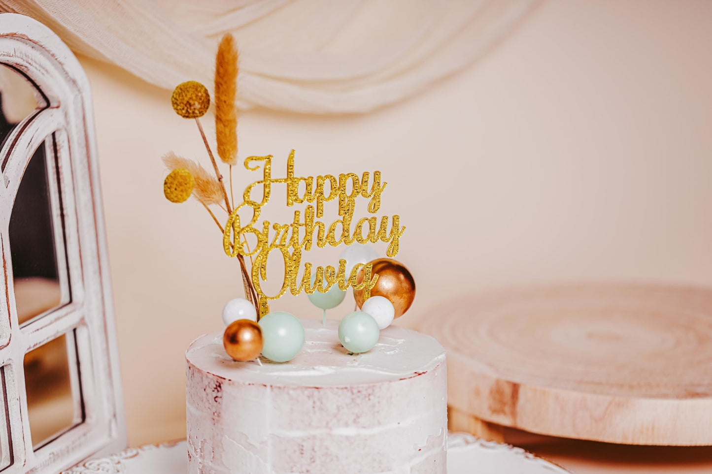 A white frosted cake featuring Mirabilia Boutique's Acrylic Happy Birthday Cake Topper, personalized with "Happy Birthday Olivia" in gold, is decorated with small green, gold, and white balls. The elegant design is beautifully complemented by a backdrop of soft draped fabric and wooden accents.