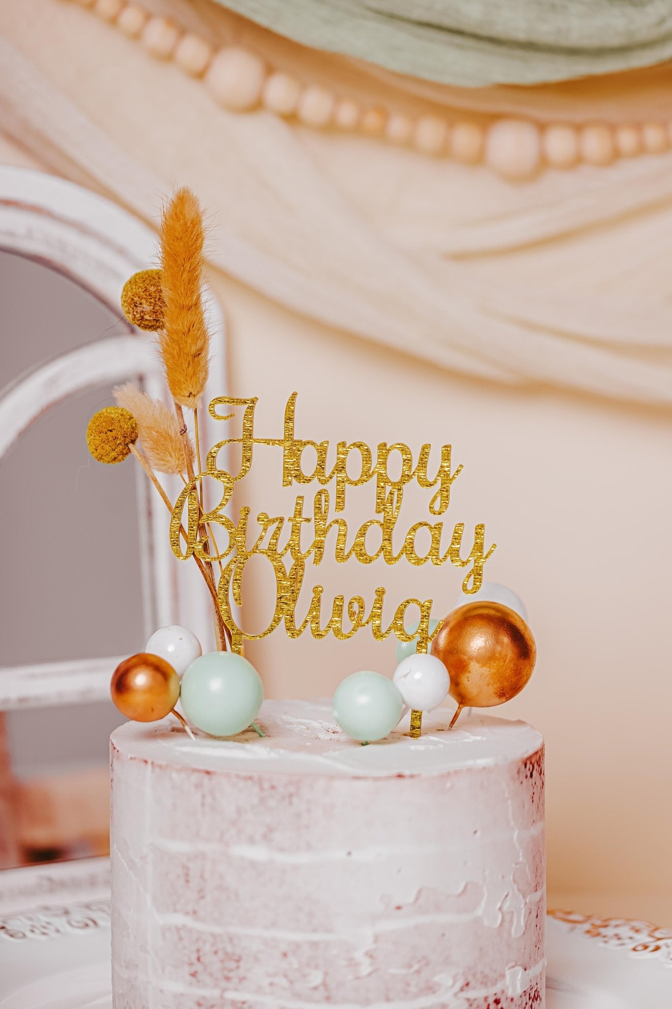 A pastel-colored birthday cake features the Mirabilia Boutique's Acrylic Happy Birthday Cake Topper for Festive Celebrations, personalized with "Olivia," and embellished with decorative spheres and dried flowers. The custom color cake is beautifully set against a soft, elegant backdrop with a white chair and draped ribbon accents.