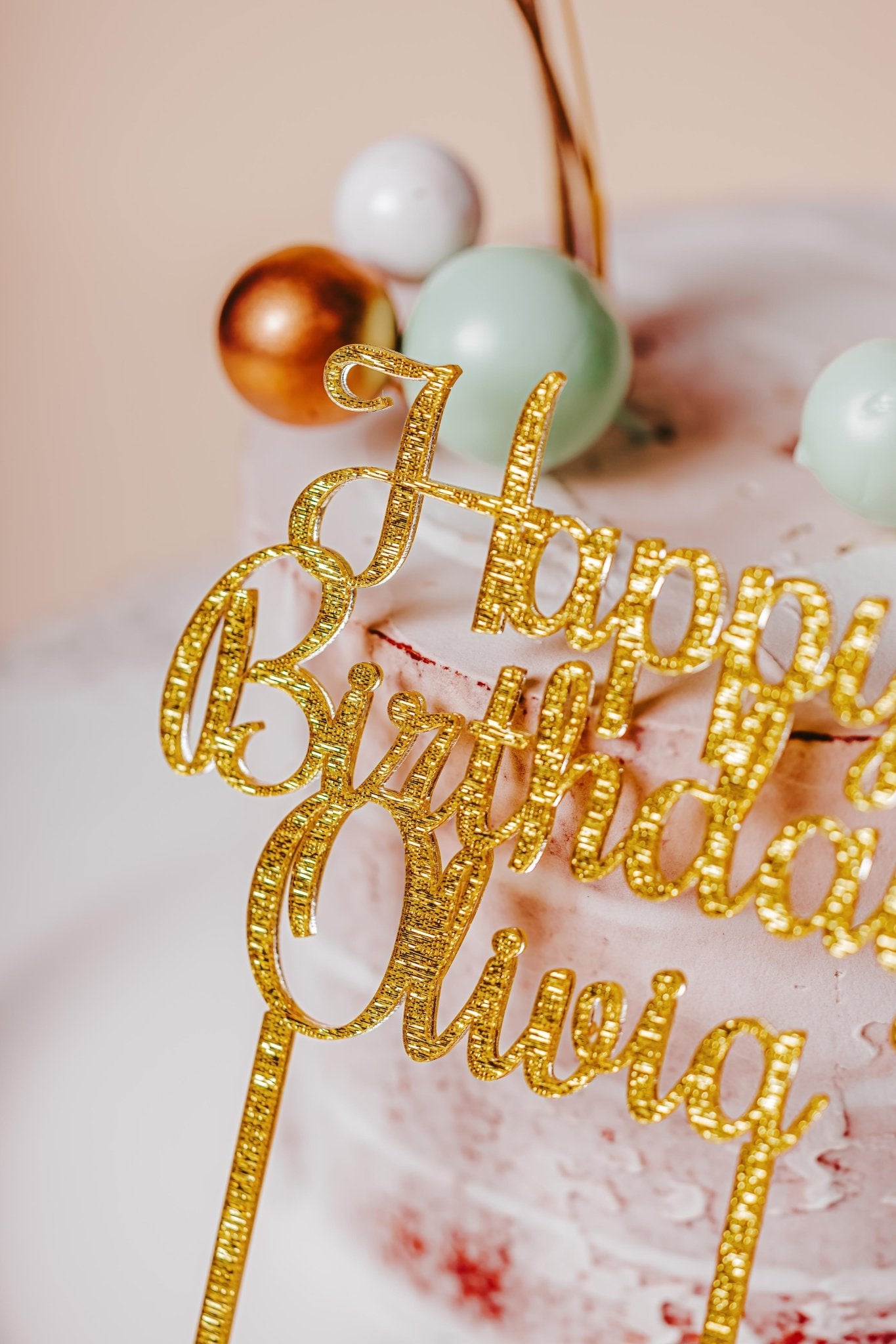 A close-up of a birthday cake featuring the Mirabilia Boutique's golden acrylic "Happy Birthday" topper. The cake is frosted with light pink icing and adorned with spherical ornaments in custom colors of white, gold, and mint green.