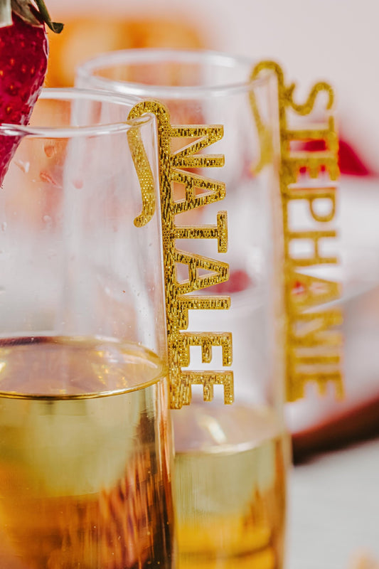 Two champagne flutes brimming with a golden beverage feature Mirabilia Boutique's Custom Acrylic Wine Glass Names for Events & Celebrations. The tag on the front flute displays "NATALEE," while the one behind reads "STEPHANIE." A strawberry playfully rests on the rim of one glass, enhancing its charm.