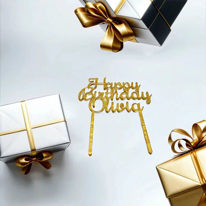 Three gift boxes wrapped in silver paper with gold ribbons are arranged around a golden Mirabilia Boutique Acrylic Happy Birthday Cake Topper on a white background.