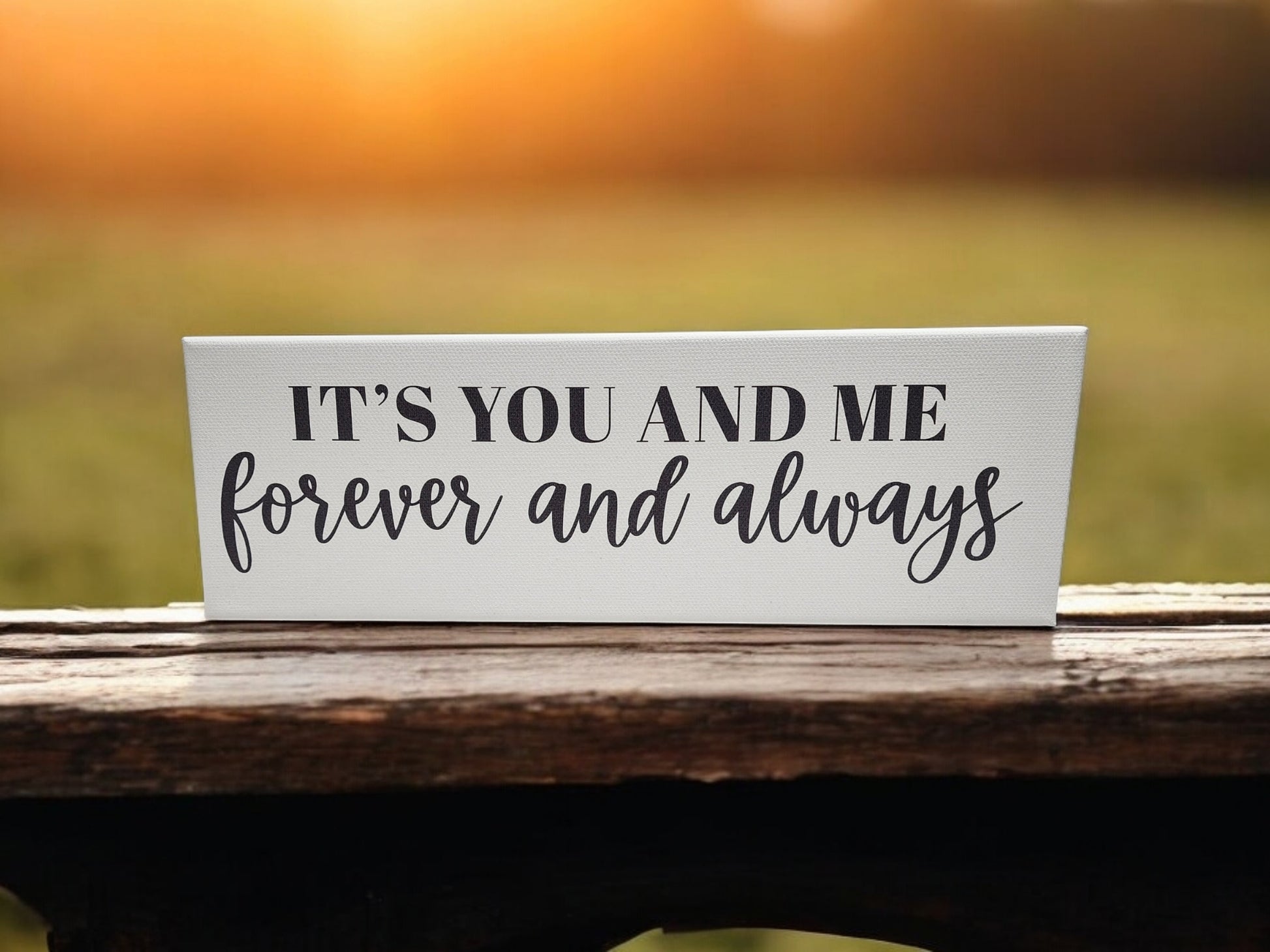 The "It's You and Me Forever and Always." 4x12 Cradled Canvas by Mirabilia Boutique features black typography on wood, resting on a wooden surface. Framed by a warm, blurred sunset background, this romantic wall art creates an inviting atmosphere.