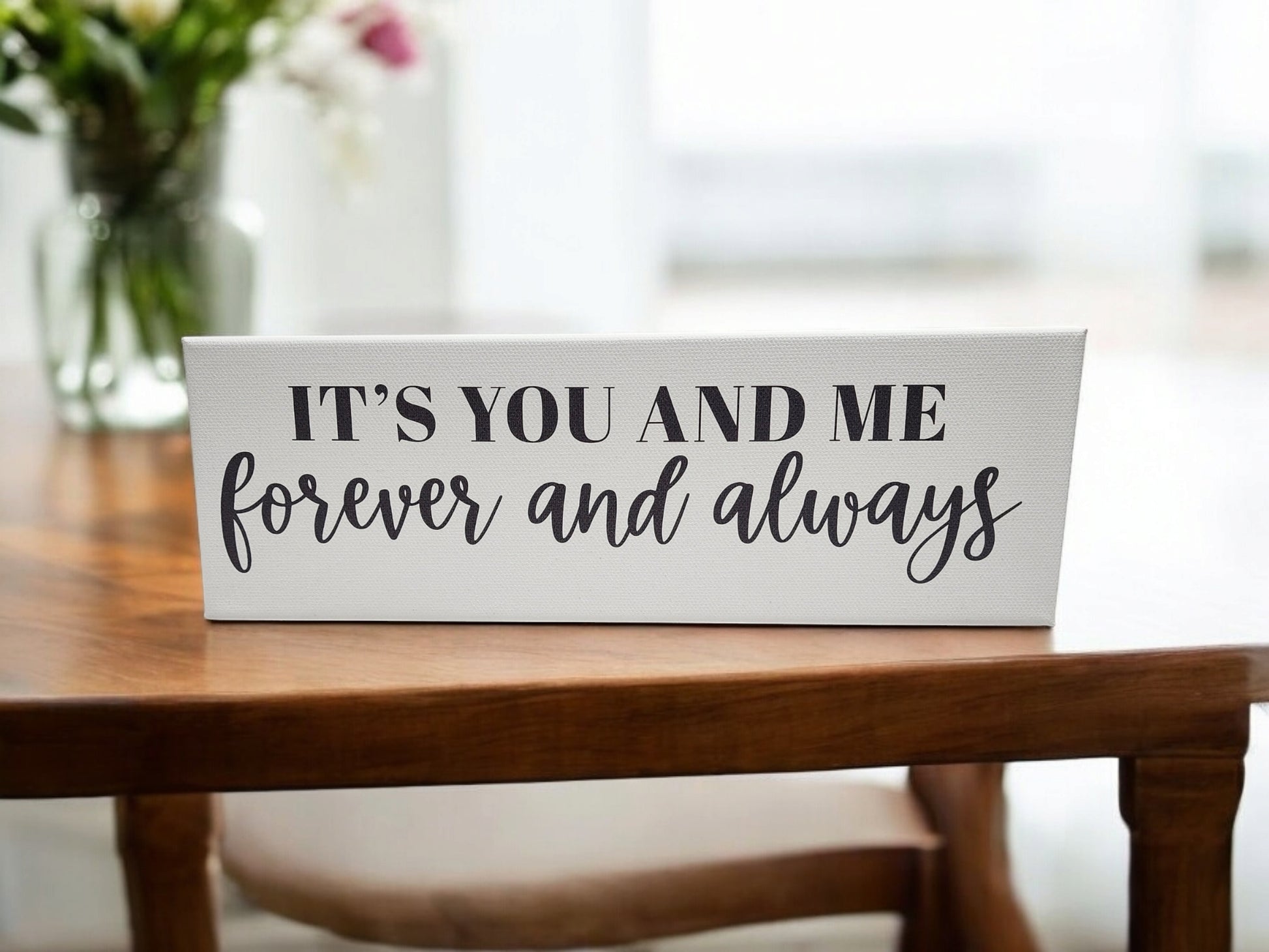 A 4x12 white cradled canvas from Mirabilia Boutique reads "It's You and Me Forever and Always" in bold black typography on a wooden table, with a blurred vase of flowers in the background adding charm to this piece of personalized home decor.