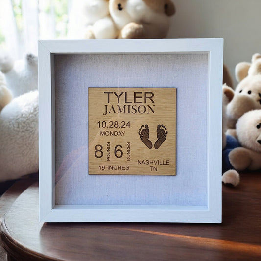 Mirabilia Boutique's Baby Birth Stats Nursery Sign displays intricately engraved details: "Tyler Jamison, 10.28.24, Monday, 8 pounds, 6 ounces, 19 inches, Nashville, TN," complete with footprints. In the background of this personalized keepsake are blurred plush toys.