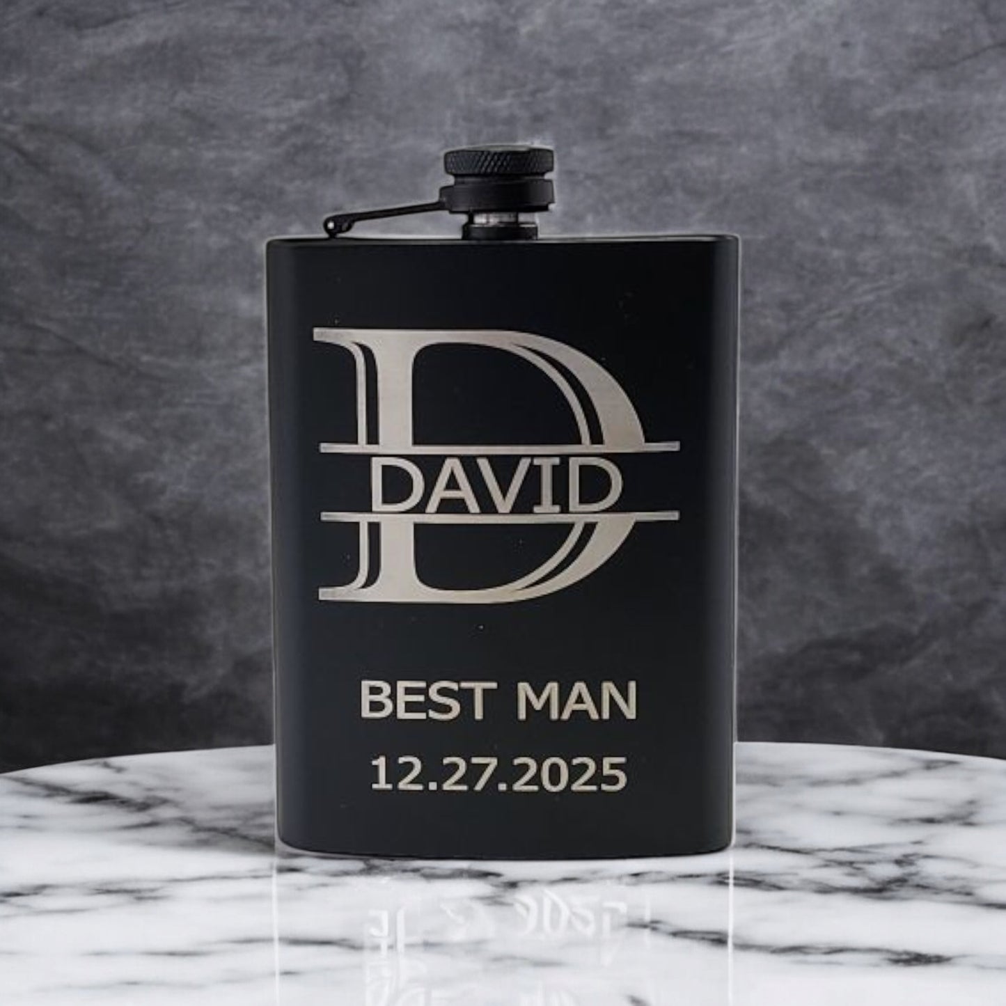 This Personalized Matte Black Hip Flask by Mirabilia Boutique features custom engraving with "D," "David," "Best Man," and the date "12.27.2025." It rests elegantly on white marble against a dark, textured backdrop. Perfect as a custom gift for him!.