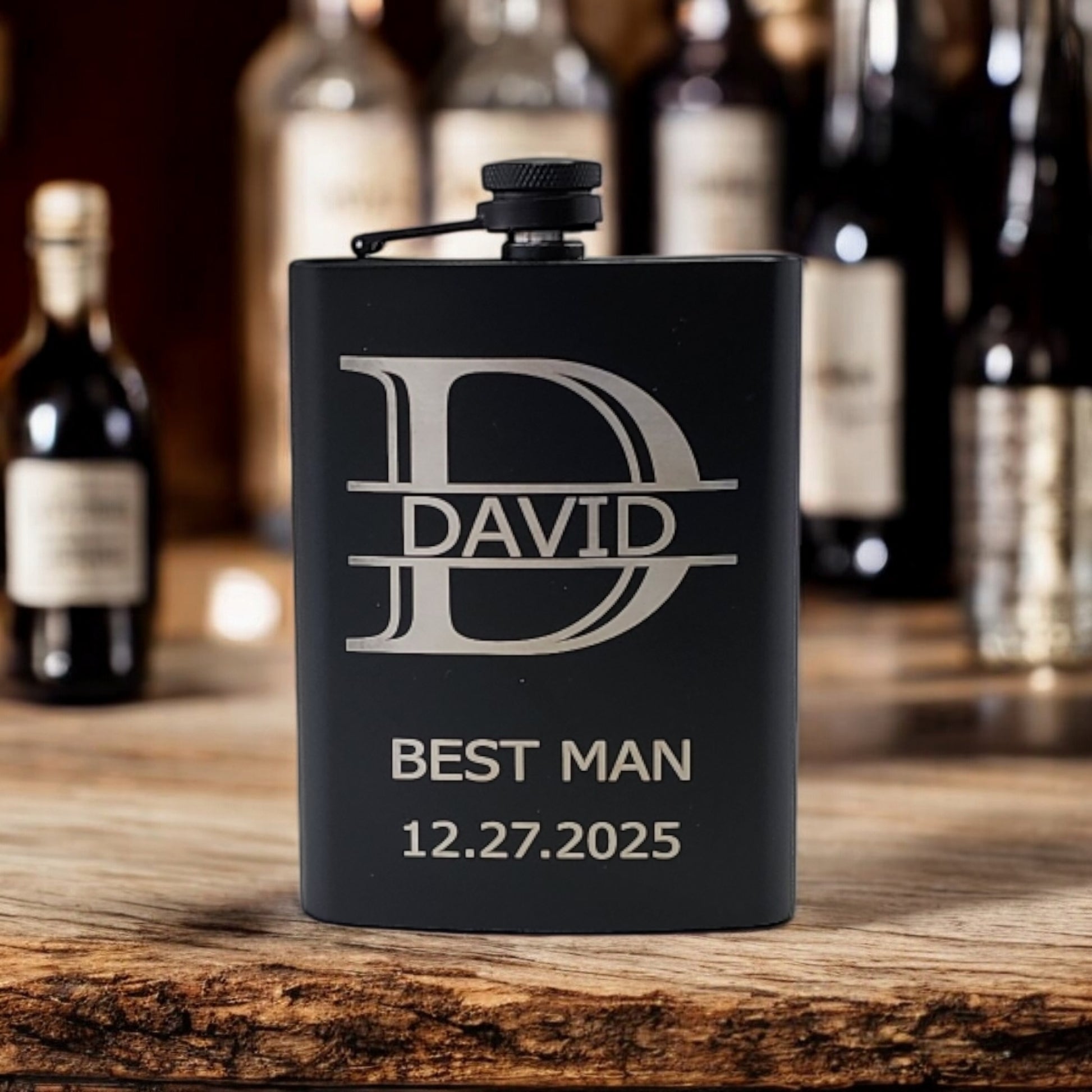 A Personalized Matte Black Hip Flask by Mirabilia Boutique lies on a wooden surface, engraved with "David," "Best Man," and "12.27.2025." Blurred bottles in the background hint at a bar setting, highlighting the elegance of this custom gift for him.