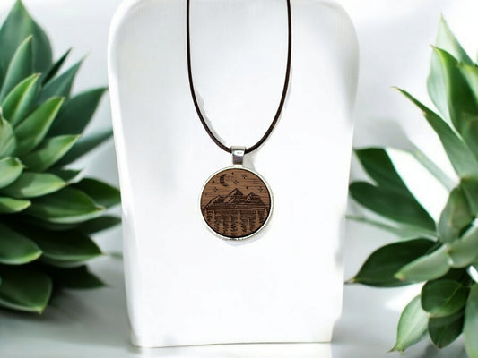 The Walnut Wood Mountain Pendant Necklace by Mirabilia Boutique features a nature-inspired etched mountain and tree design, hanging on a dark cord. Displayed elegantly on a white stand, it's surrounded by lush green succulent plants.