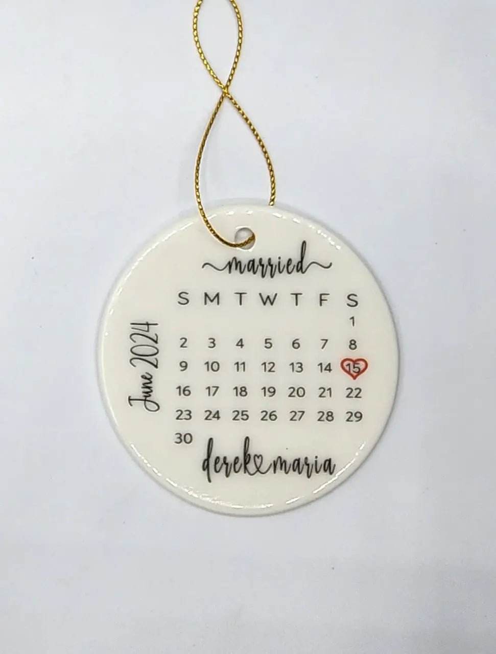 This custom personalized ceramic wedding ornament from Mirabilia Boutique showcases a round design with a June 2014 calendar. The 15th is beautifully highlighted with a heart, and the words "married" and "derek & maria" are elegantly displayed on its surface. It features a gold loop for convenient hanging, making it an ideal personalized keepsake.