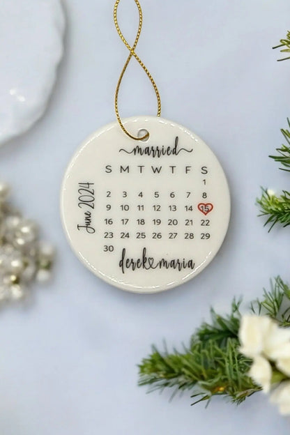 The Custom Personalized Ceramic Wedding Ornament for Couples, crafted by Mirabilia Boutique, showcases a June 2014 calendar with "married" elegantly inscribed at the top. The date 21 is highlighted with a red heart, and beneath it, "derek & maria" is beautifully written in cursive. Enhanced with a gold string and encircled by greenery, this ornament serves as a stunning custom wedding keepsake.