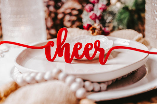 A holiday dining setup features Mirabilia Boutique's Festive Christmas Plate Words for Holiday Cheer, with the word "cheer" elegantly scripted in red across its surface. The background is embellished with holiday decor, incorporating touches of green, red, and natural textures to evoke a warm and inviting festive ambiance.