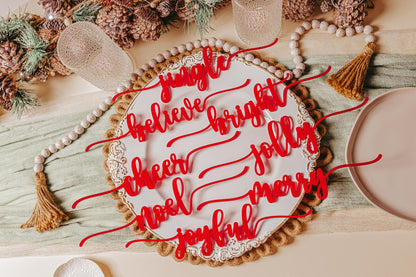 The Festive Christmas Plate Words for Holiday Cheer by Mirabilia Boutique is a round white plate embellished with joyful words like "jingle," "believe," and "merry" in red, arranged in a circular pattern. It's adorned with pinecones, greenery, and decorative tassels, making it an ideal addition to your holiday tablescape.