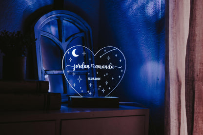 A Personalized Wedding Date Night Light Custom Keepsake from Mirabilia Boutique glows softly on a bedside table, featuring the names "Jordan" and "Amanda" along with the date "12.28.2023." The illuminated heart showcases moon and star designs that cast a gentle blue light in a dimly lit room, making it an ideal personalized wedding gift and memorable wedding date keepsake.