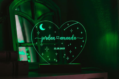 A heart-shaped LED light from Mirabilia Boutique's Personalized Wedding Date Night Light Custom Keepsake collection glows green, displaying the names "Jordan" and "Amanda", with the date "12.28.2025" beneath. This personalized wedding gift is adorned with stars and a crescent moon, positioned in front of a reflective surface featuring a vintage design in the background.