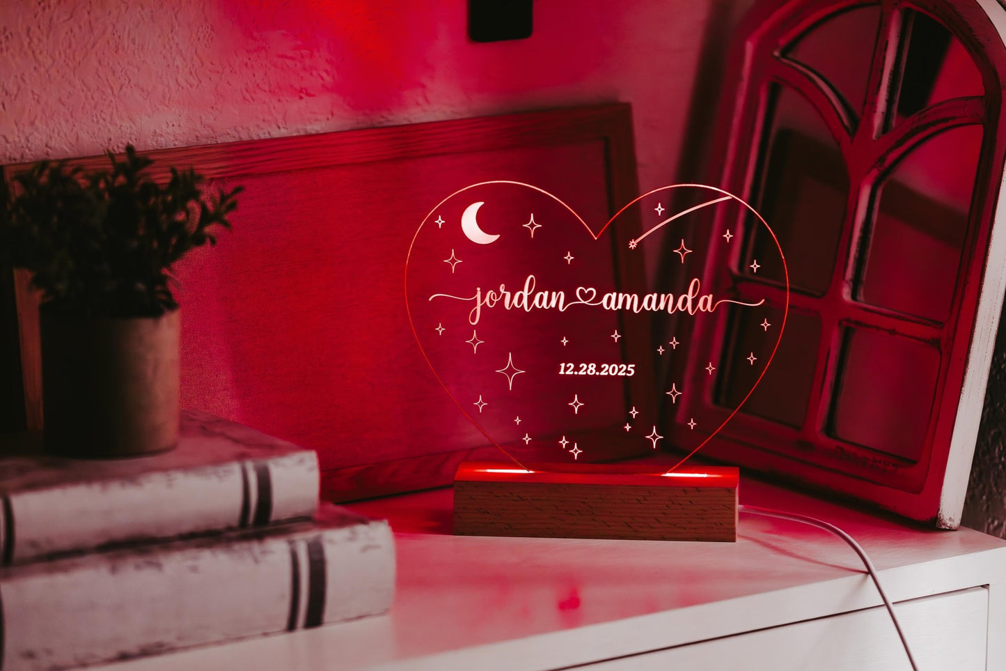 The Mirabilia Boutique Personalized Wedding Date Night Light Custom Keepsake features a heart-shaped acrylic design mounted on a wooden base. It elegantly displays the names "Jordan" and "Amanda" along with their wedding date, "12.28.2025." This customized wedding gift emits a warm red glow and is surrounded by various decor items such as books, a small plant, and a window-shaped mirror.