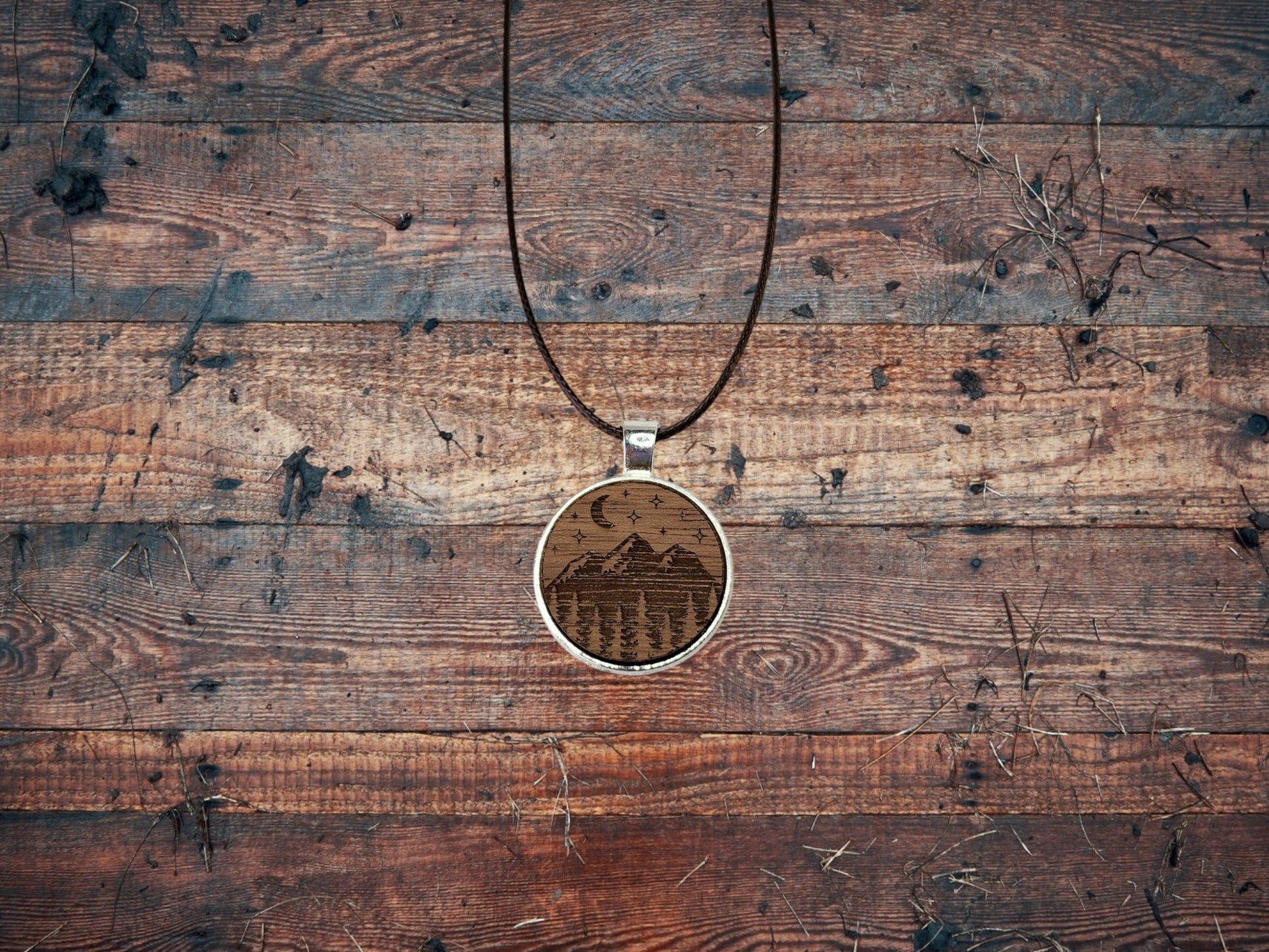 The Walnut Wood Mountain Pendant Necklace by Mirabilia Boutique features a crescent moon and stars on a circular pendant, hanging from a thin leather cord. Resting on rustic walnut wood, it epitomizes nature-inspired jewelry with its natural and earthy feel.