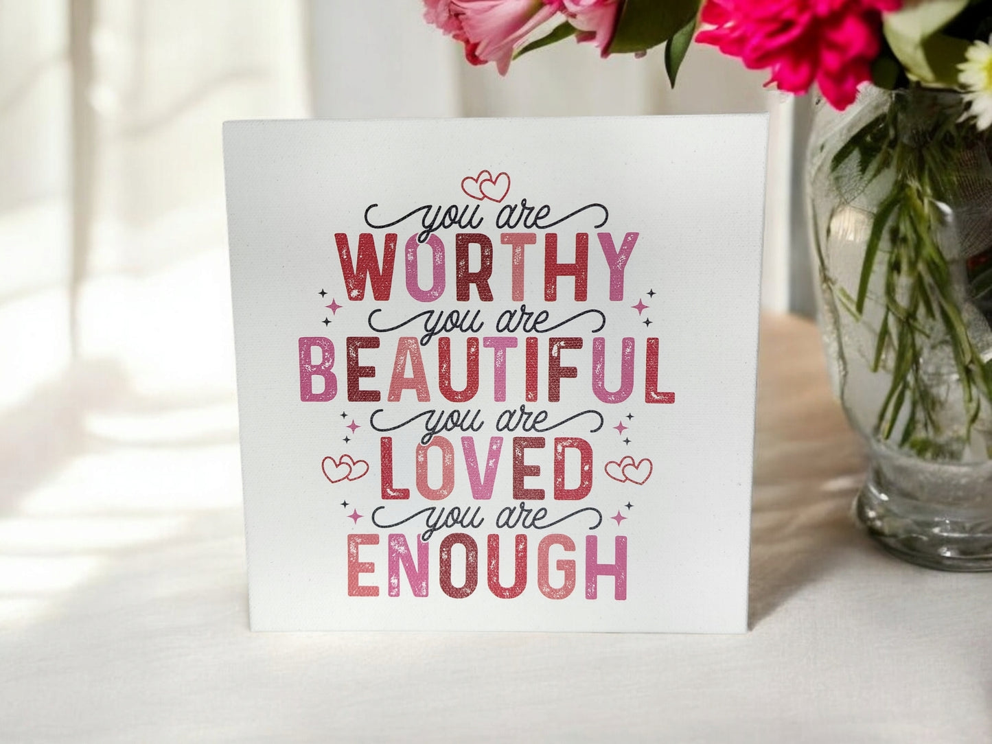 The 8x8 "You Are Worthy, Beautiful, Loved, Enough" Cradled Canvas Sign by Mirabilia Boutique features pink and red text with heart designs. It serves as an empowering affirmation surrounded by a vase with pink flowers to brighten any space.