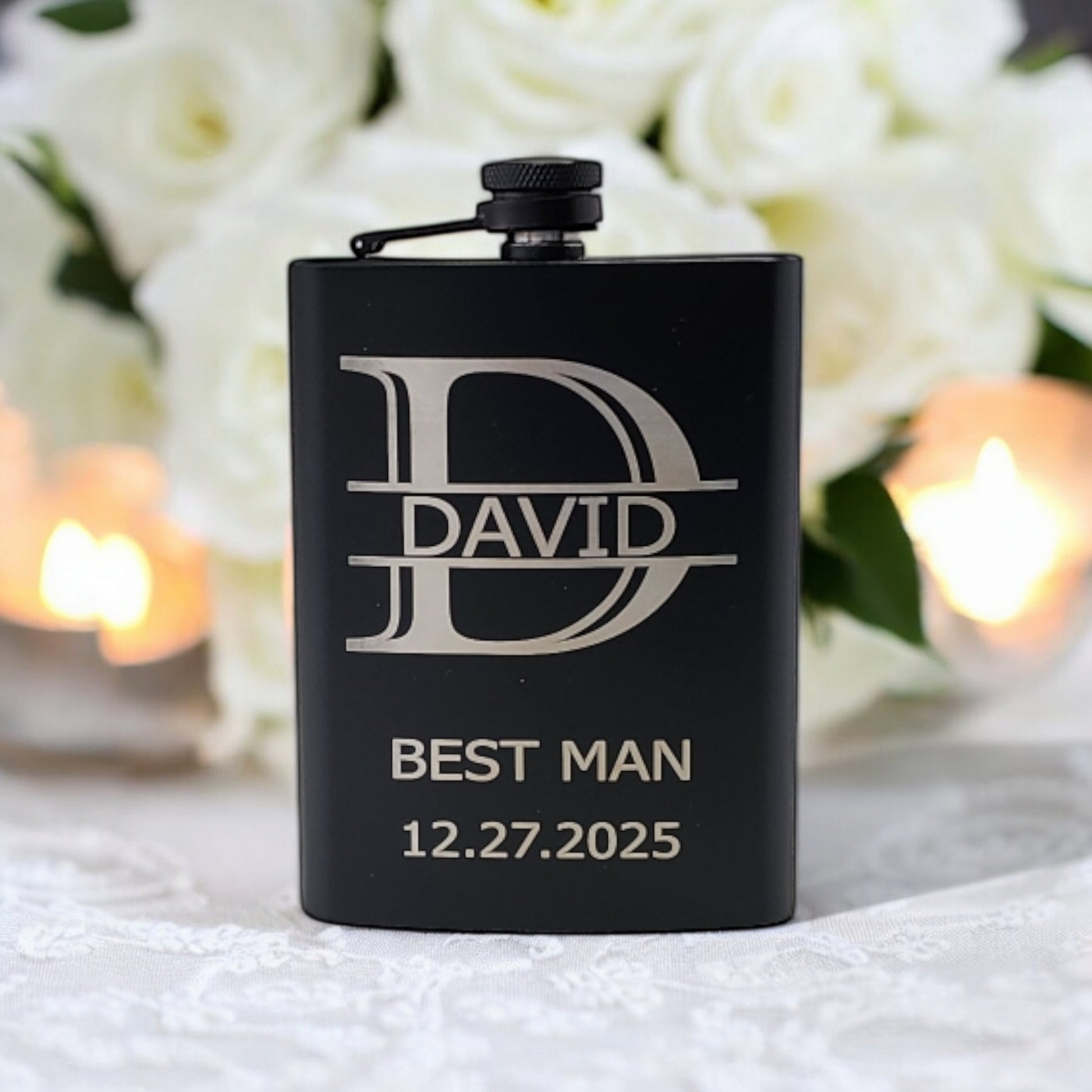 A Matte Black Hip Flask engraved with "David, Best Man, 12.27.2025" from Mirabilia Boutique elegantly rests on lace, with soft-focused white roses and lit candles enhancing the romantic ambiance.