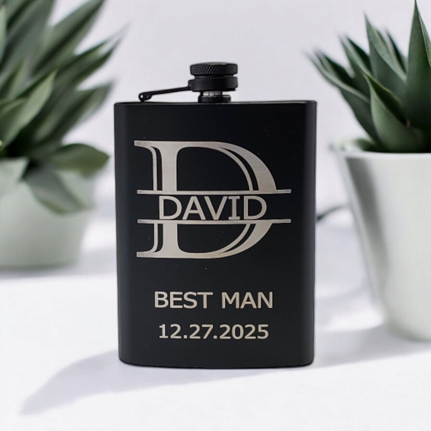 A Mirabilia Boutique matte black hip flask, personalized with "David" in a large stylized font, "Best Man," and the date "12.27.2025," is flanked by two small green plants in white pots.