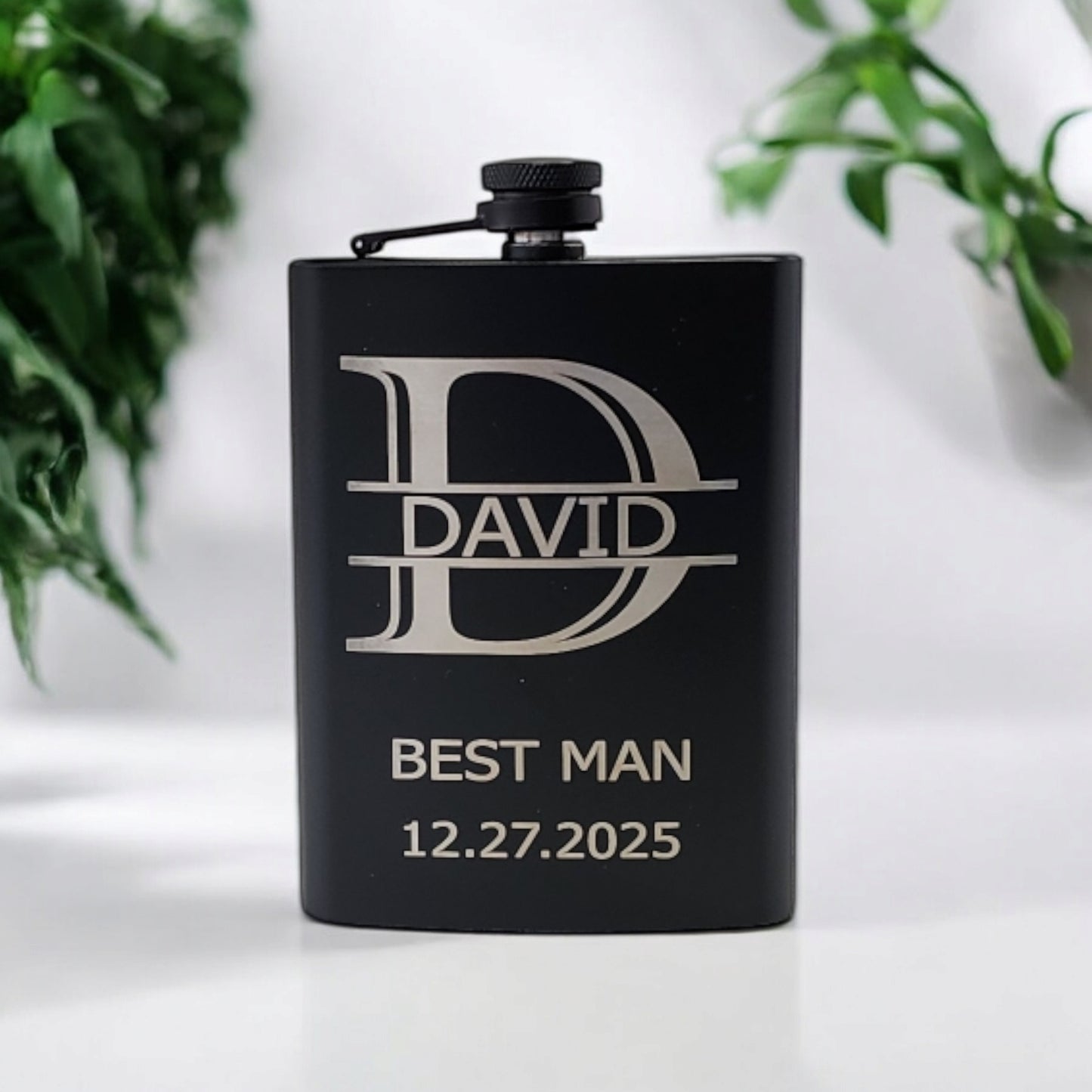 The Mirabilia Boutique Personalized Matte Black Hip Flask features "DAVID" and "BEST MAN 12.27.2025" elegantly engraved for a sleek touch, with two blurred plants in the background.