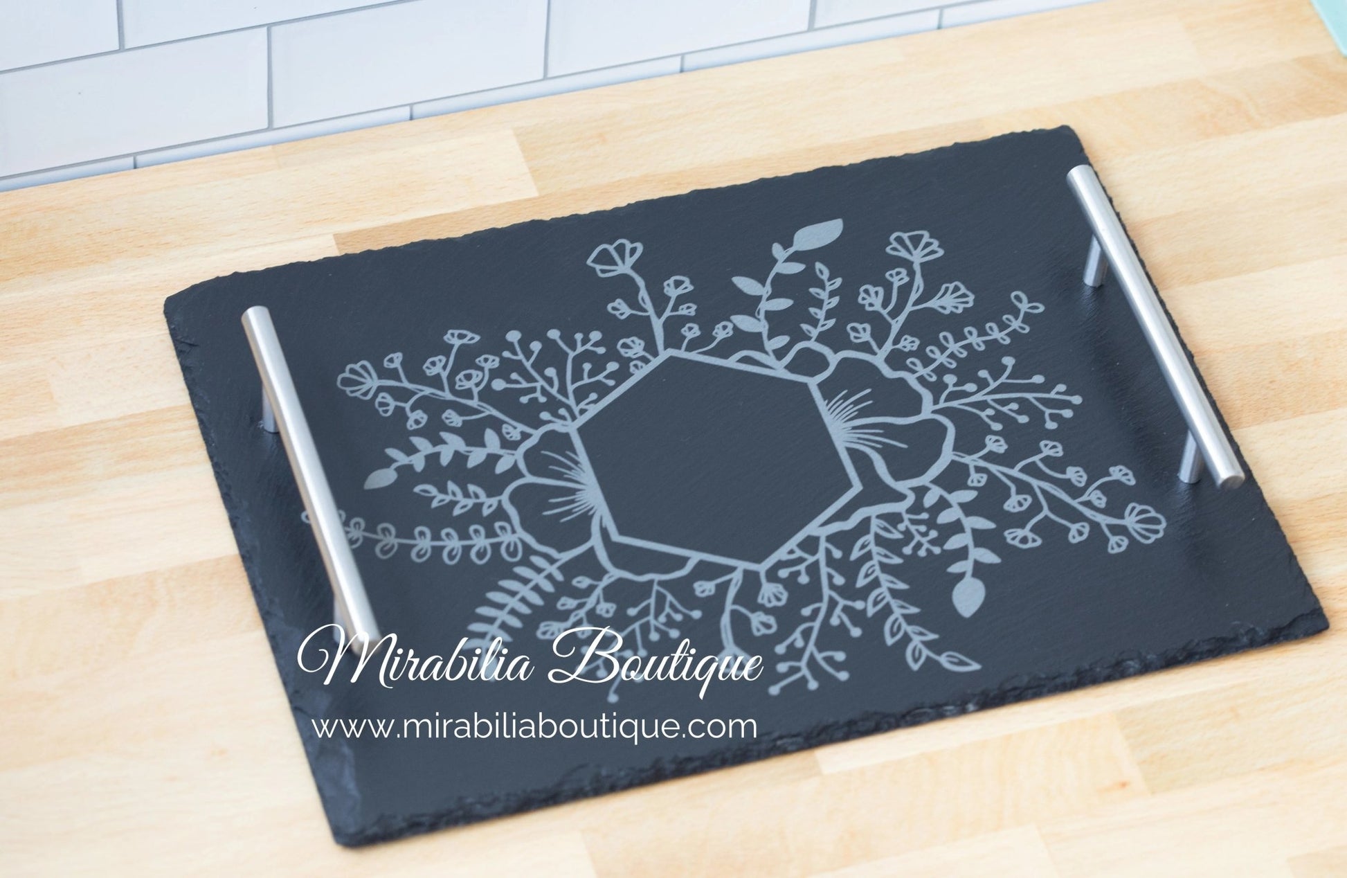 The Personalized Floral Engraved Slate Tray from Mirabilia Boutique highlights a geometric floral design etched in the center and features silver handles. The text "Mirabilia Boutique" and the website "www.mirabiliaboutique.com" are elegantly showcased on this charming piece, resting gracefully on a wooden surface.