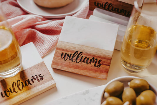 A custom engraved marble and acacia coaster set featuring the name "Williams" is tastefully arranged alongside three bottles of beverage and a bowl of olives, set against a pink cloth backdrop. The display exudes sophistication, ideal for highlighting your unique style with personalized decor from Mirabilia Boutique.