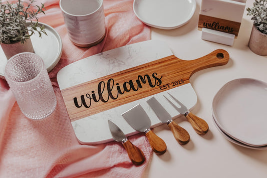 Introducing a personalized keepsake, the "Williams EST. 2025" engraved Marble and Acacia serving board by Mirabilia Boutique, designed for a perfect charcuterie setup. Surrounded by wooden-handled cheese knives, plates, a glass, and potted plants on a pink textured cloth, it’s ideal for any gathering.