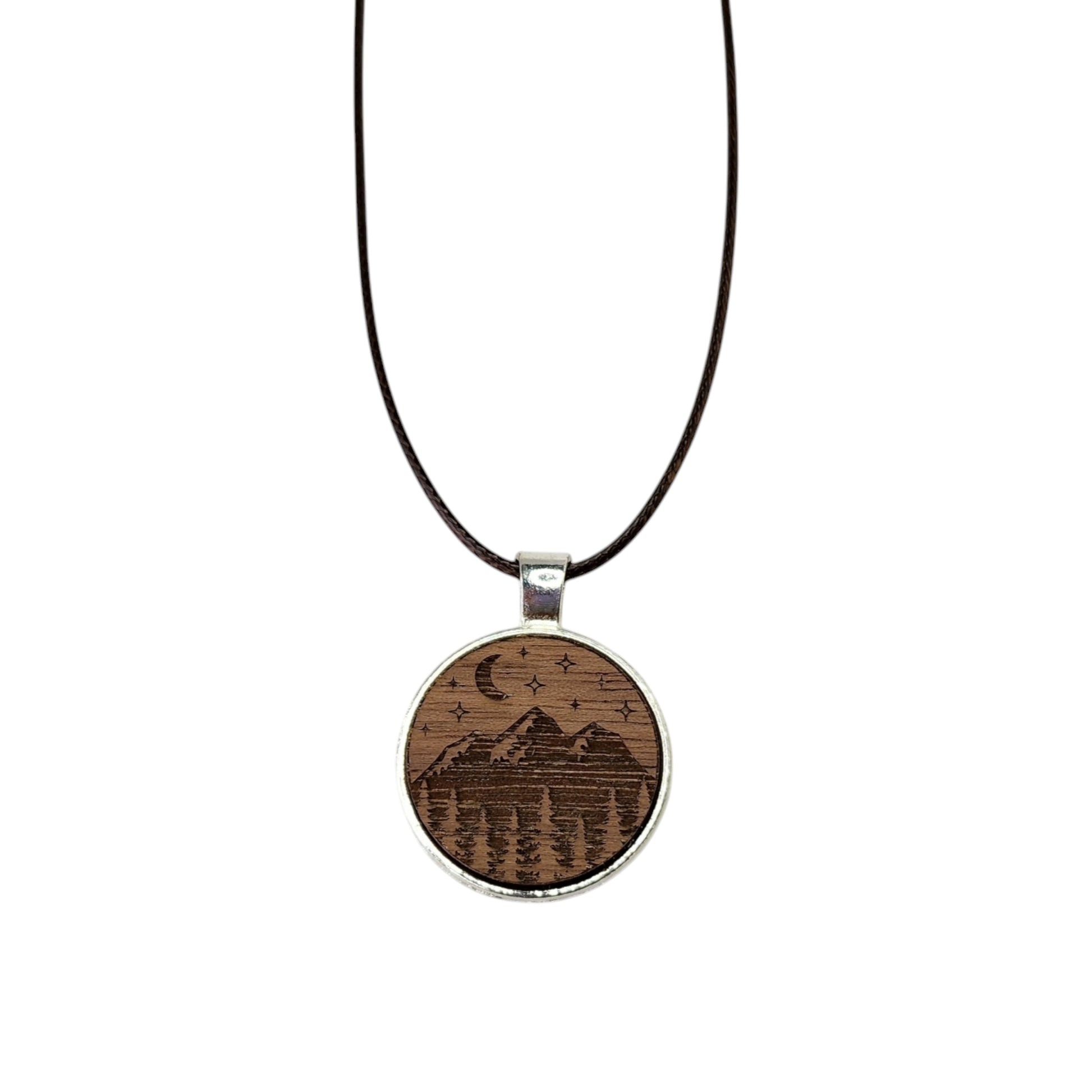 The Mirabilia Boutique Walnut Wood Mountain Pendant Necklace features a circular pendant with a mountain scene, crescent moon, and stars engraving, set in a silver-colored frame and hung on a black cord.