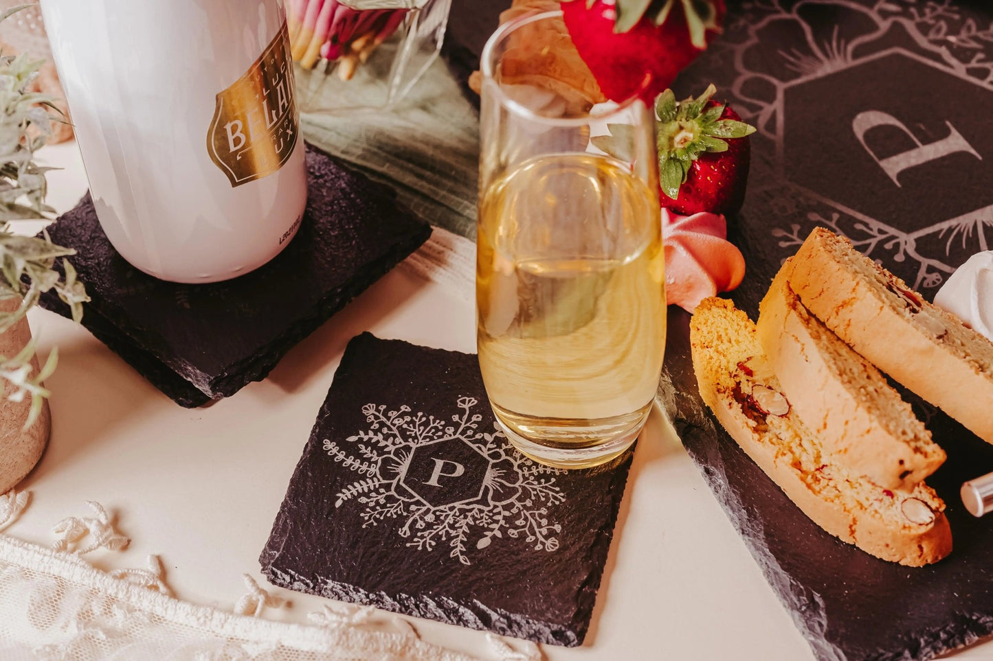 A table displays a glass of white wine perched on a Personalized Engraved Slate Coaster with a floral design and the letter "P" from Mirabilia Boutique. Nearby, biscotti, strawberries, and a white mug are placed on another coaster monogrammed with a "B." The scene is elegantly framed by plants and decorative elements.