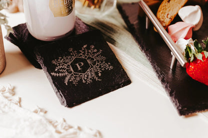 A Personalized Engraved Slate Coaster from Mirabilia Boutique, showcasing a monogram letter "P" encompassed by detailed floral motifs, rests beside a cup and a plate full of assorted desserts like strawberries and pastries on a white surface.