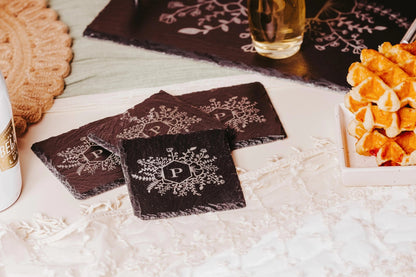 A cozy table setting with Mirabilia Boutique's Personalized Engraved Slate Coasters, showcasing a floral design and the letter "P." A stack of waffles and a beverage in a glass nearby complement the setup, all placed on a lace tablecloth for added personalized charm.