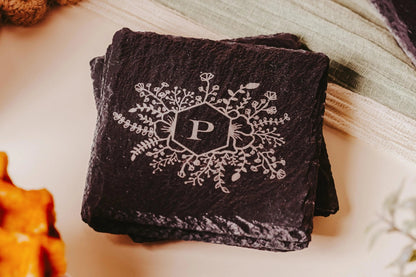 A stack of Mirabilia Boutique's Personalised Engraved Slate Coasters for Any Occasion, featuring a decorative white floral pattern surrounding a custom-engraved monogram "P" in the center, rests on a light-colored surface with soft fabric in the background.