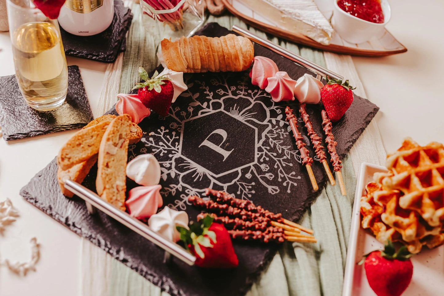 A chic charcuterie board with a central "P" monogram is artfully displayed on Mirabilia Boutique's Personalized Floral Engraved Slate Tray for Unique Gifts, featuring elegant silver handles. Encircled by strawberries, pastries, breadsticks, meringues, and waffles, the presentation is completed with wine and cheese gracefully placed in the background.