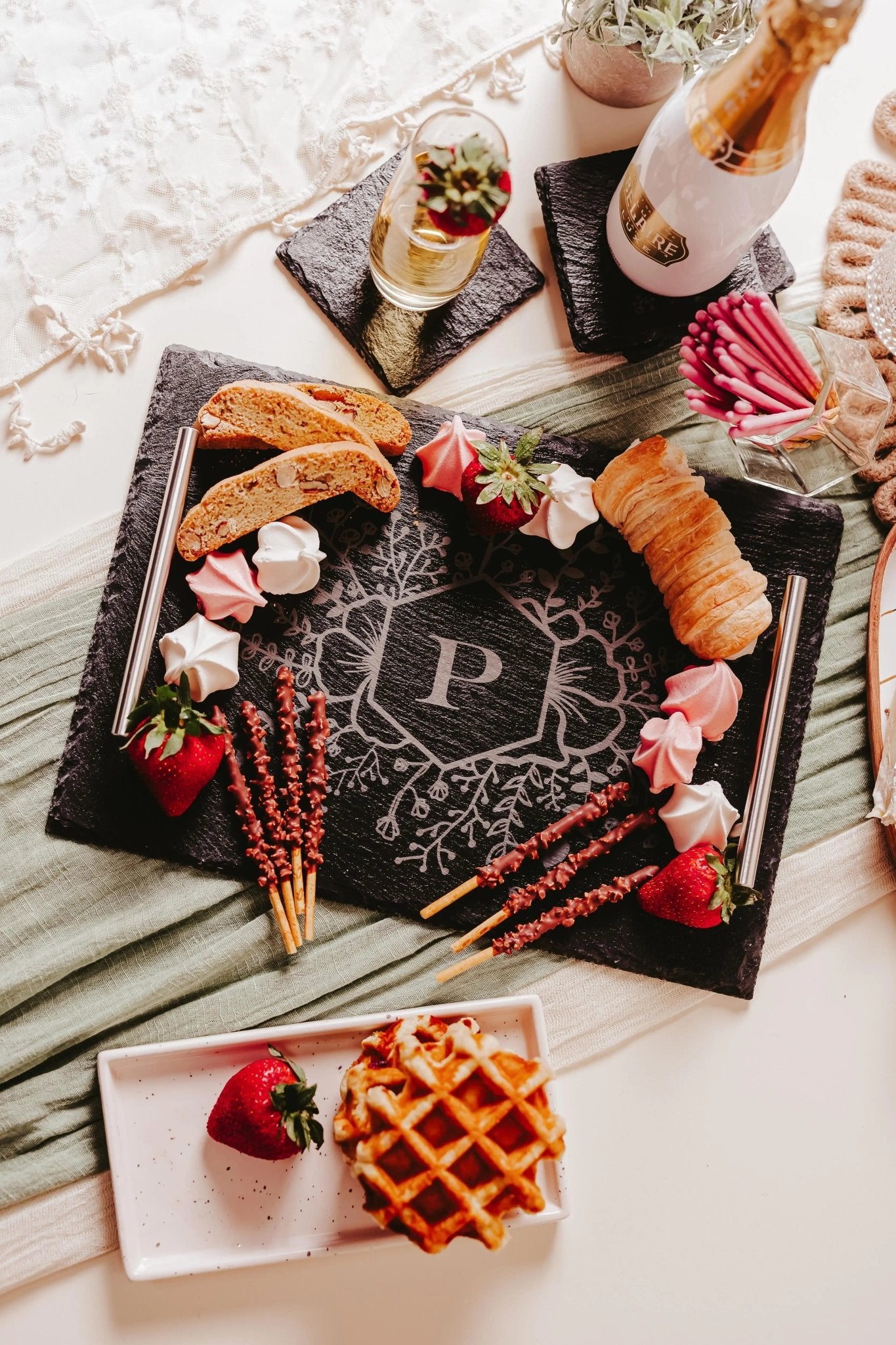 The Personalized Floral Engraved Slate Tray for Unique Gifts by Mirabilia Boutique showcases a delightful floral motif with the letter 'P' at its center. It is beautifully arranged with strawberries, meringues, biscotti, bread, and pretzel sticks. An elegant setting is completed with a plate featuring a waffle and strawberry alongside a bottle of champagne in the background.