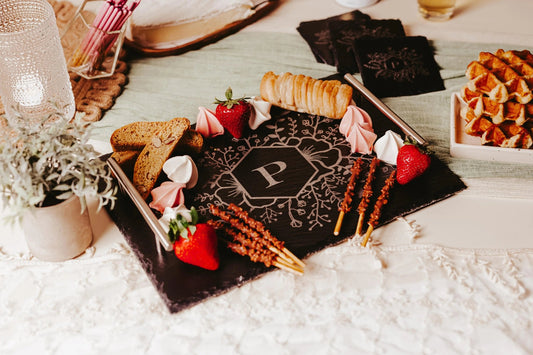 A Personalized Floral Engraved Slate Tray by Mirabilia Boutique, featuring a monogram "P" and silver handles, is surrounded by strawberries, pastries, biscotti, pink and white meringues, and pretzel sticks. A bowl of flowers sits on the left side, while waffles are placed on a plate to the right, all set against a lace tablecloth.