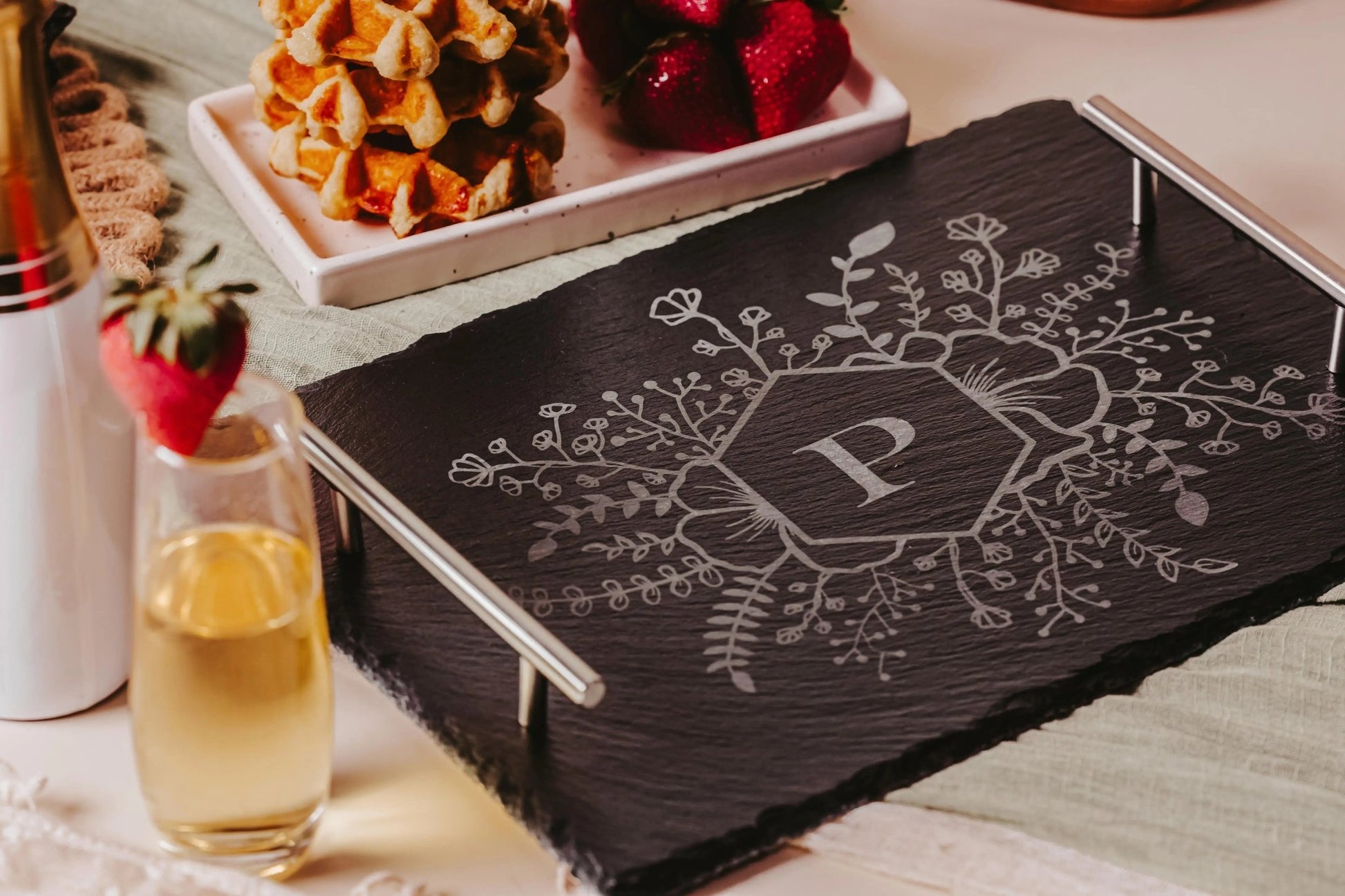 The Personalized Floral Engraved Slate Tray for Unique Gifts, from Mirabilia Boutique, features intricate floral designs and a central engraved letter "P." It is elegantly positioned next to a champagne flute, waffles topped with strawberries, and fresh strawberries on the table.