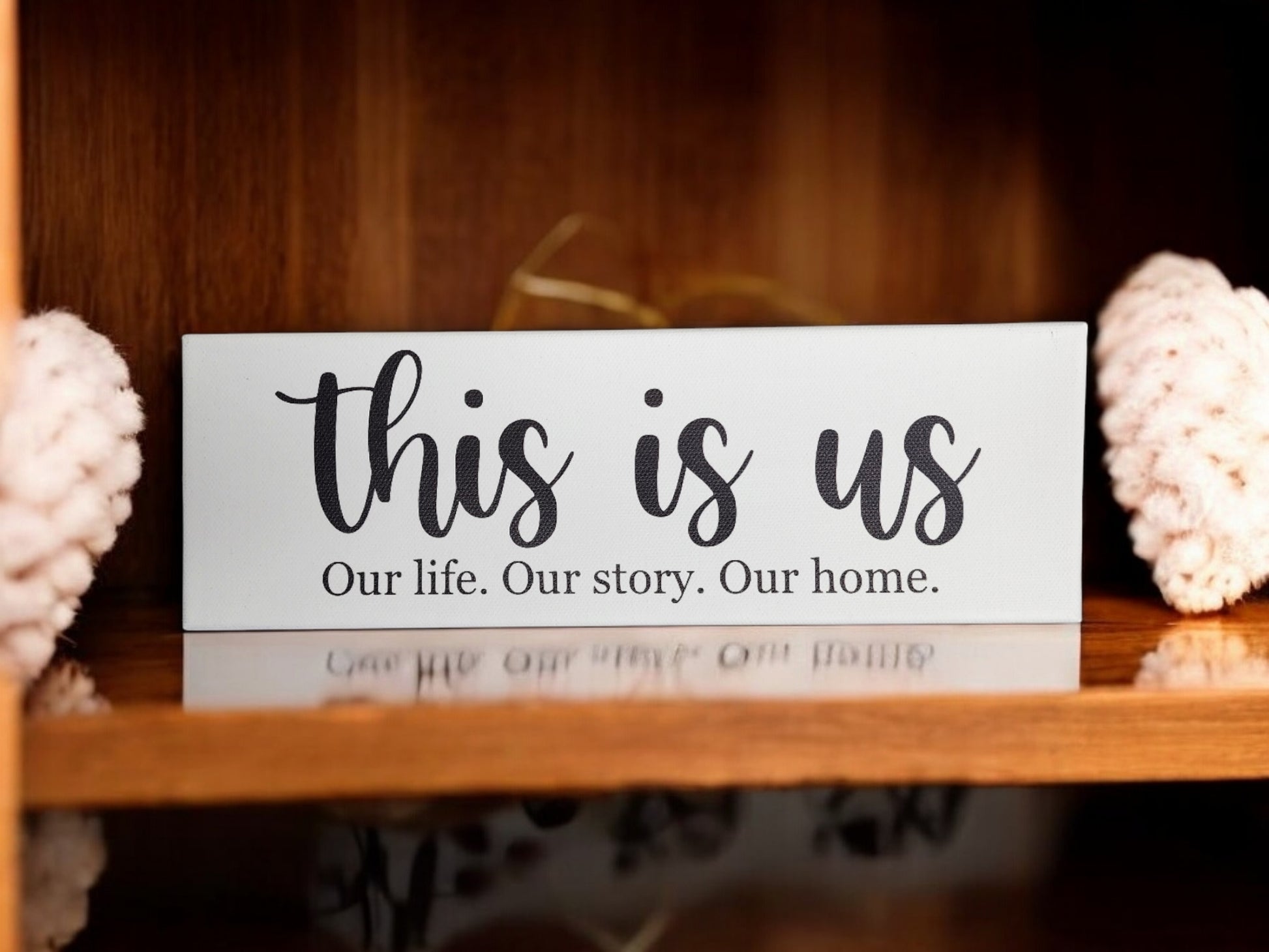 A wooden shelf displays Mirabilia Boutique's "This Is Us. Our Life. Our Story. Our Home." 4x12 Cradled Canvas, featuring elegant black script and smaller print, flanked by knitted items for a timeless decor statement.