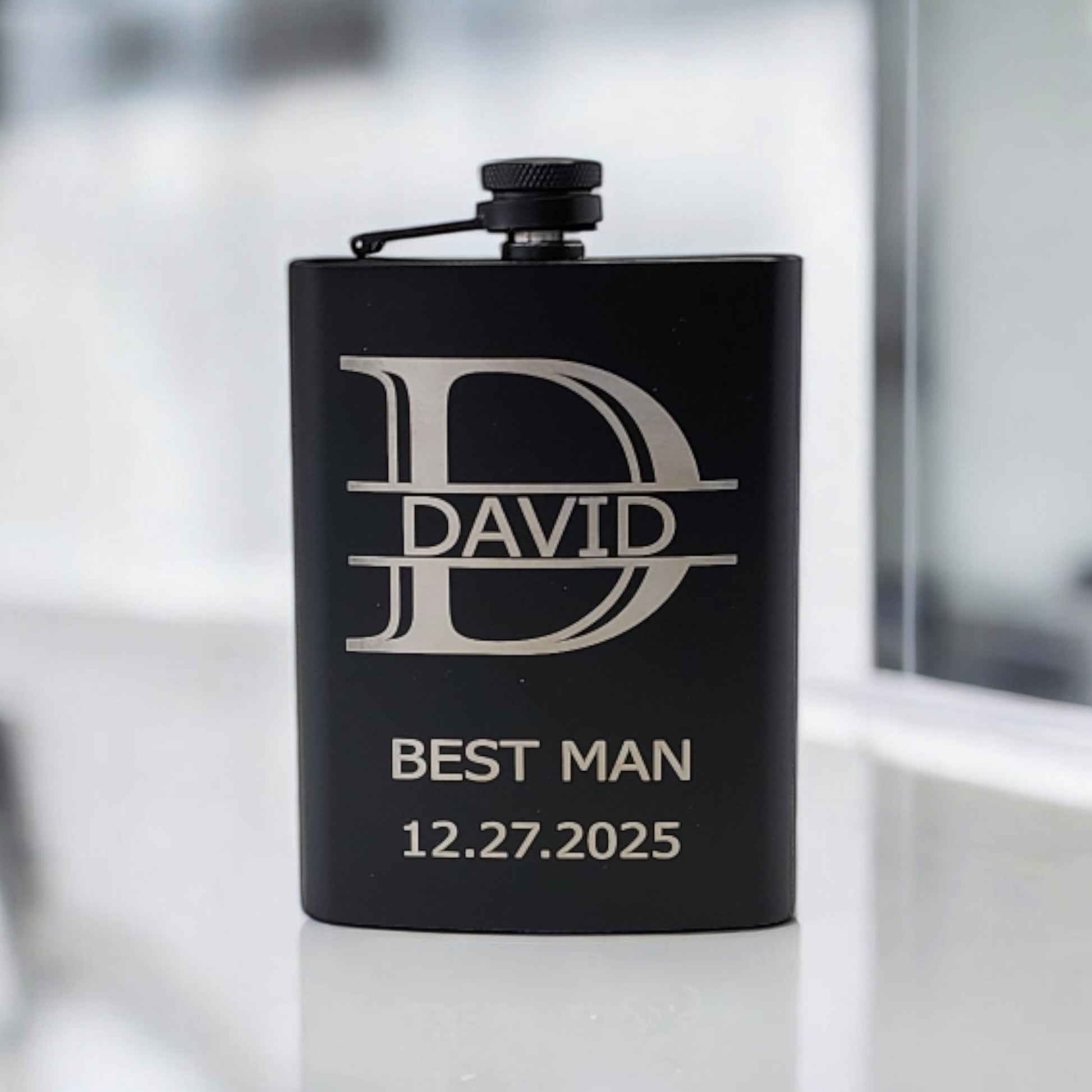 A matte black flask by Mirabilia Boutique with a silver cap rests on a reflective surface. This personalized hip flask features "DAVID" engraved inside a large "D" and "BEST MAN 12.27.2025" below, making it an ideal keepsake for special occasions.