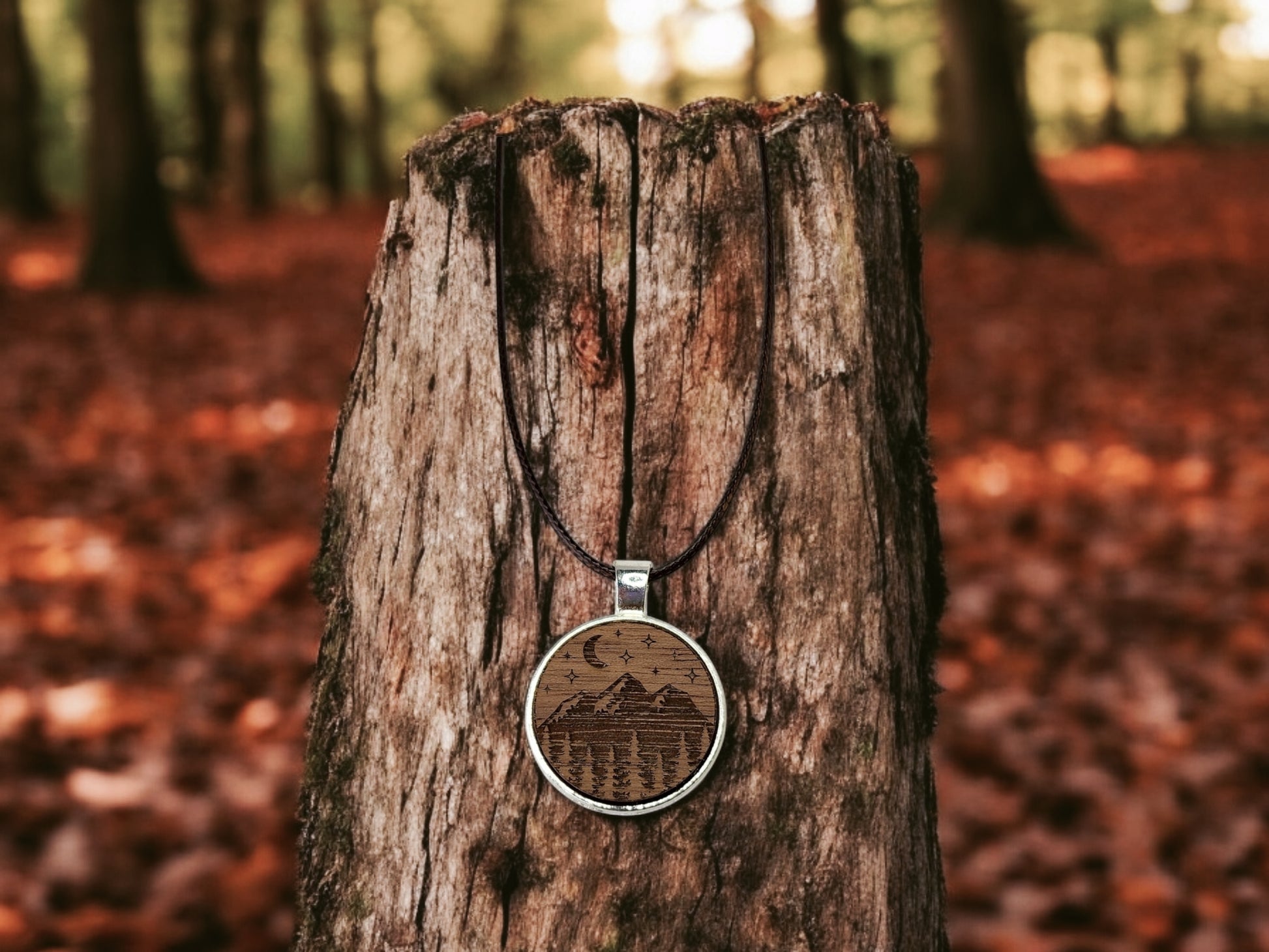 The Walnut Wood Mountain Pendant Necklace by Mirabilia Boutique features a round, nature-inspired design with an engraved mountain and forest scene. It hangs against a tree stump amidst autumn leaves, with distant trees and crescent moon & stars details.