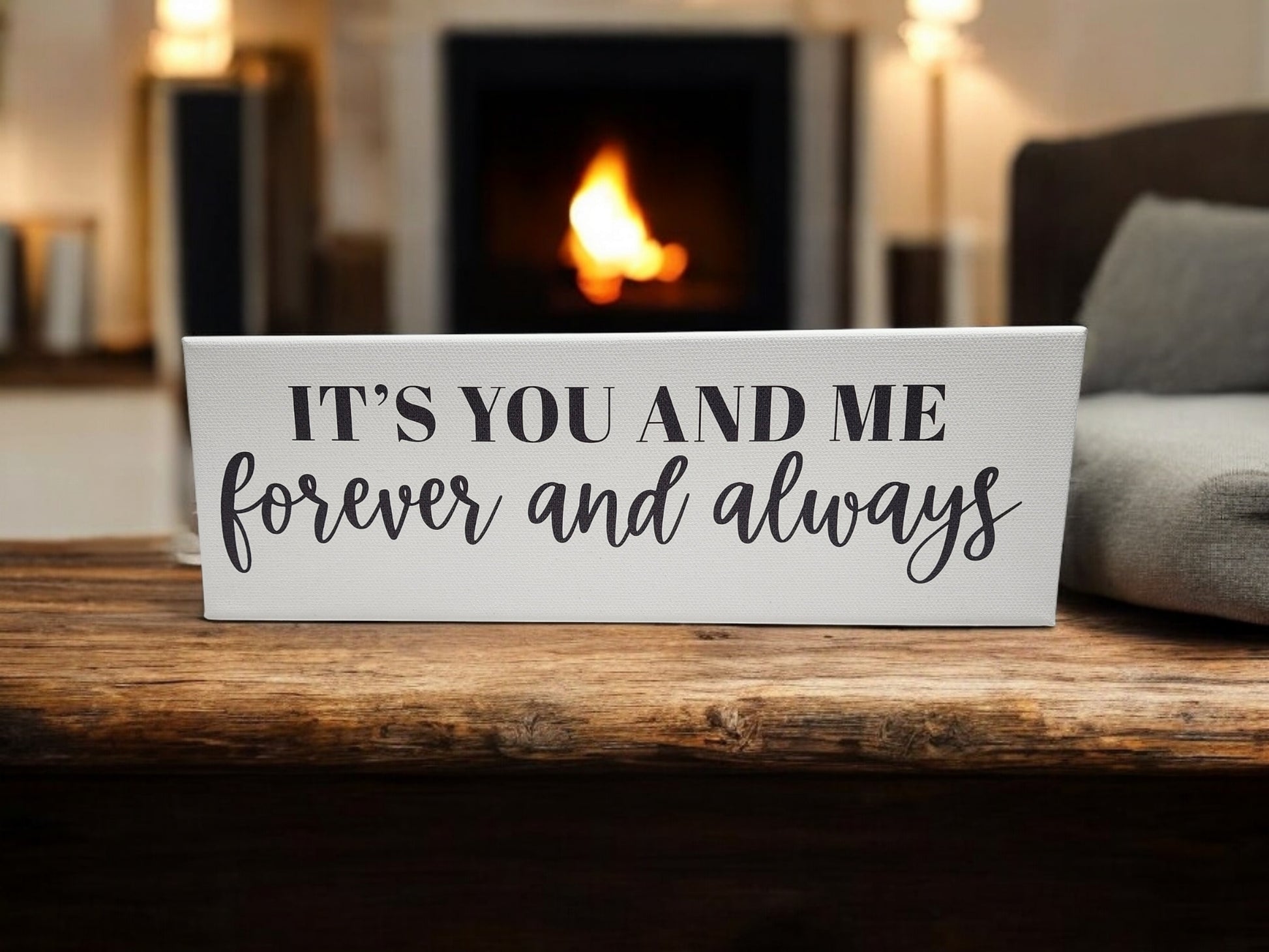 A 4x12 cradled canvas from Mirabilia Boutique reading "It's You and Me Forever and Always" rests on a wooden table. In the blurred background, a cozy fireplace adds warmth to this romantic wall art scene, enhancing its inviting ambiance.