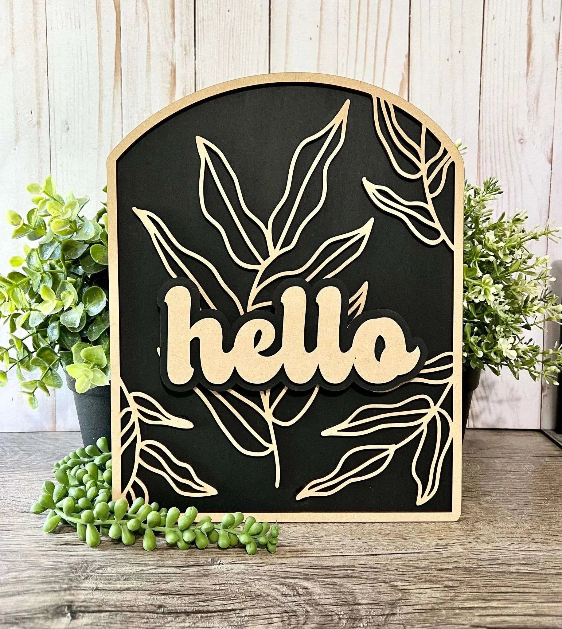 The Hello Sign Arched by Mirabilia Boutique is a charming decorative piece featuring the word "hello" in cursive font against a black background with an outline of leafy branches. Encased in a wooden frame, it rests elegantly on a wooden surface with potted green plants in the background, warmly whispering "welcome to your home.