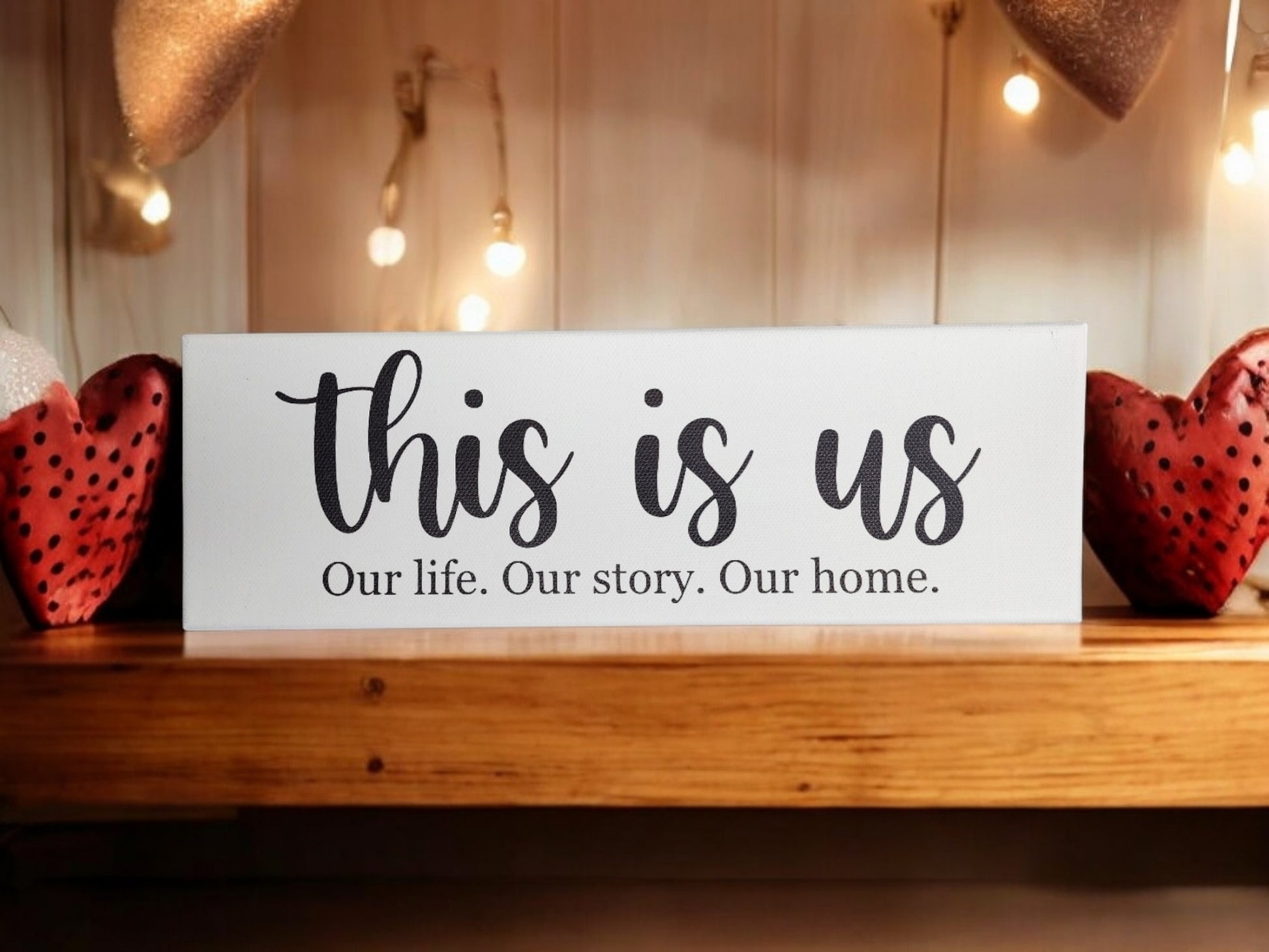 A Mirabilia Boutique 4x12 cradled canvas reading "This is us. Our life. Our story. Our home." sits on a wooden shelf between heart-shaped ornaments, creating meaningful decor. Warm string lights hang above, enhancing the timeless design.